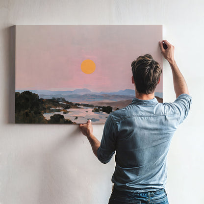 A man is hanging a large landscape painting on a wall, which depicts a serene sunset over a mountainous terrain.