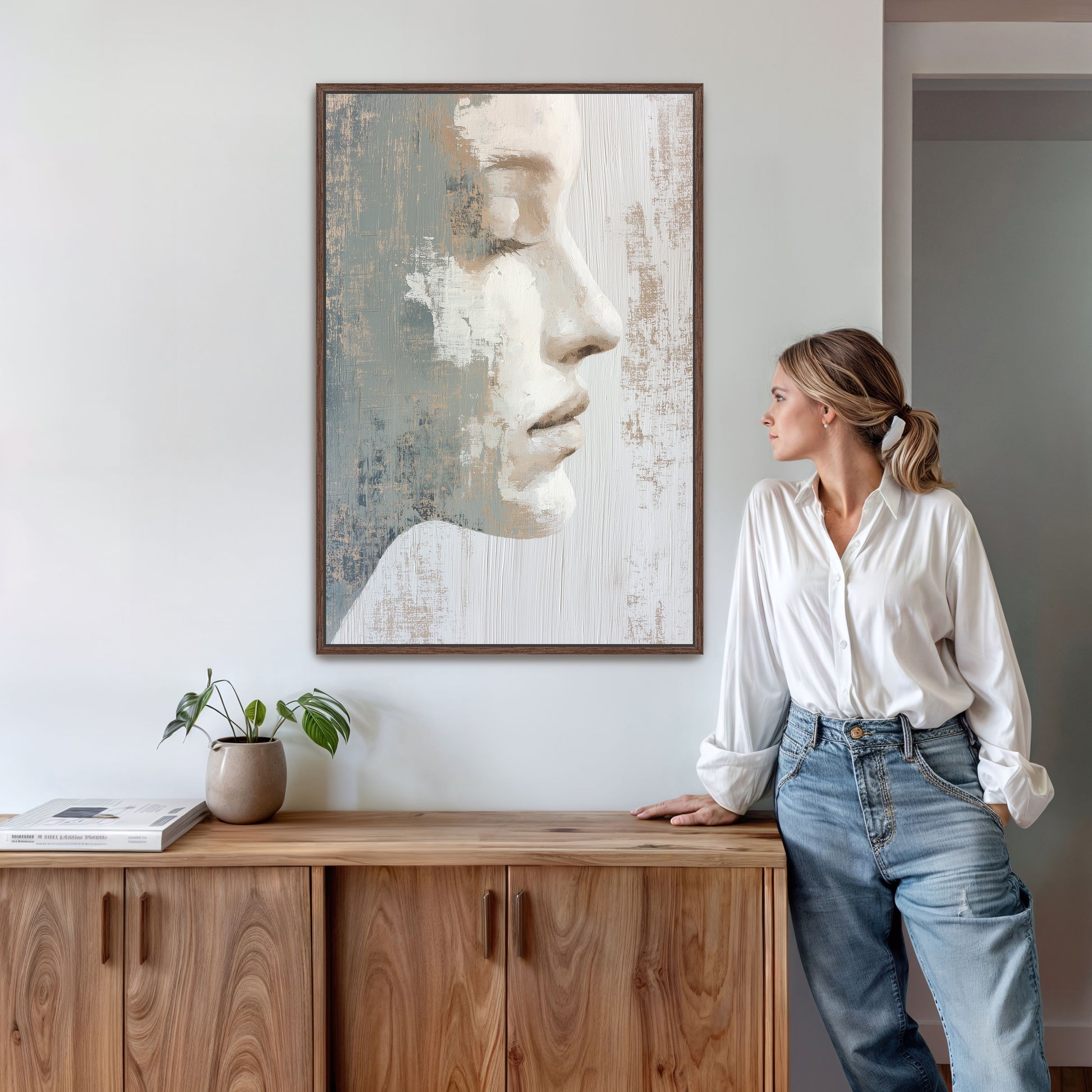 A woman in a white shirt and blue jeans stands next to a wooden cabinet, leaning against it, with a framed painting of a woman's face hanging on the wall above her.