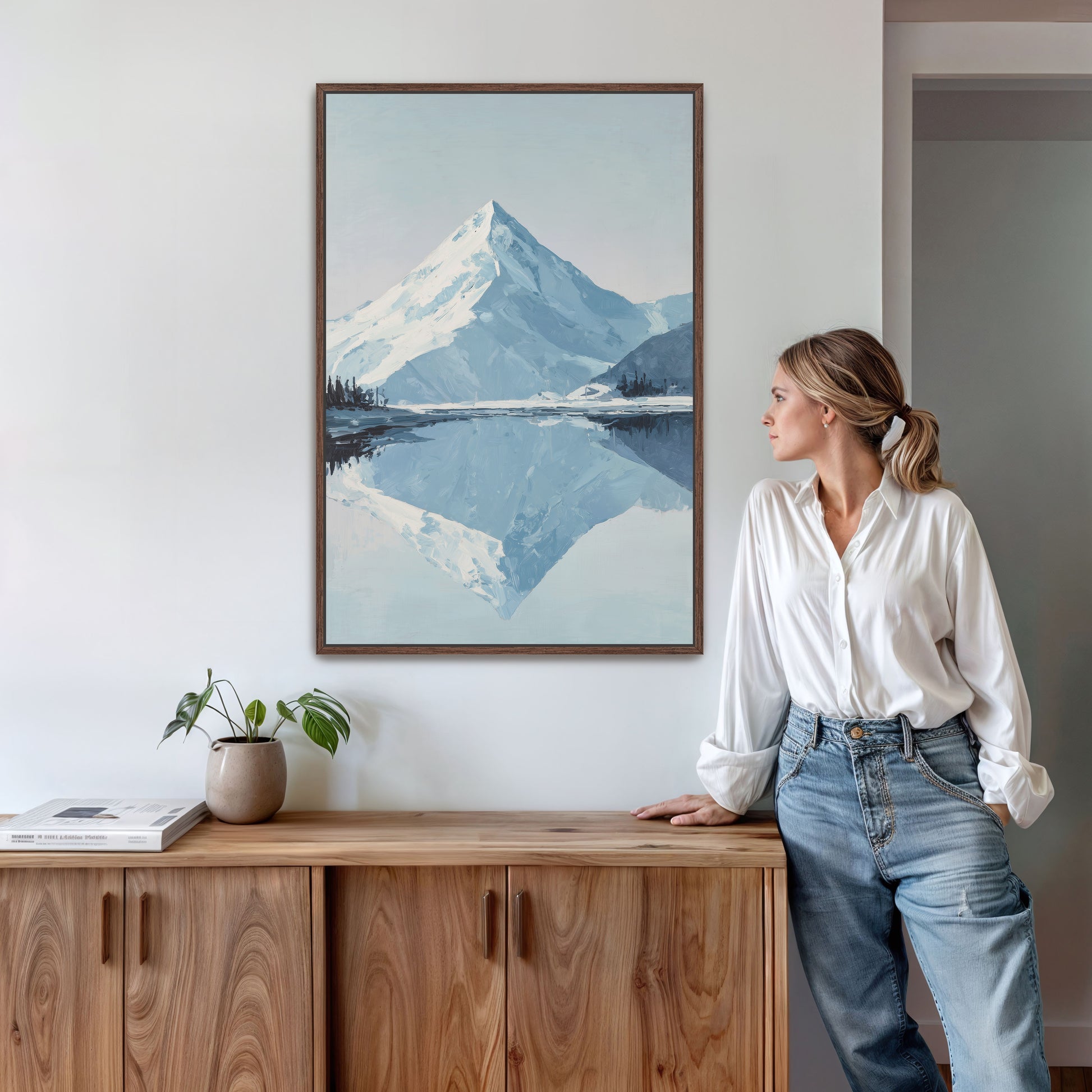 A woman in a white blouse and blue jeans stands next to a wooden cabinet, leaning against it, with a framed painting of a mountain landscape hanging on the wall above her.