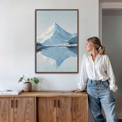 A woman in a white blouse and blue jeans stands next to a wooden cabinet, leaning against it, with a framed painting of a mountain landscape hanging on the wall above her.