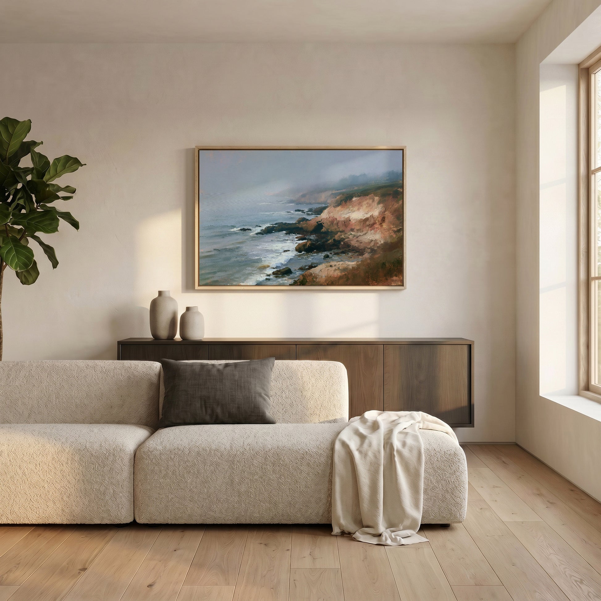 a serene living room with a large, framed painting of a coastal landscape hanging on the wall above a beige sofa. The sofa is adorned with a gray throw blanket and a white vase, adding a touch of elegance to the space. The room is illuminated by natural light streaming in from a window, creating a warm and inviting atmosphere.
