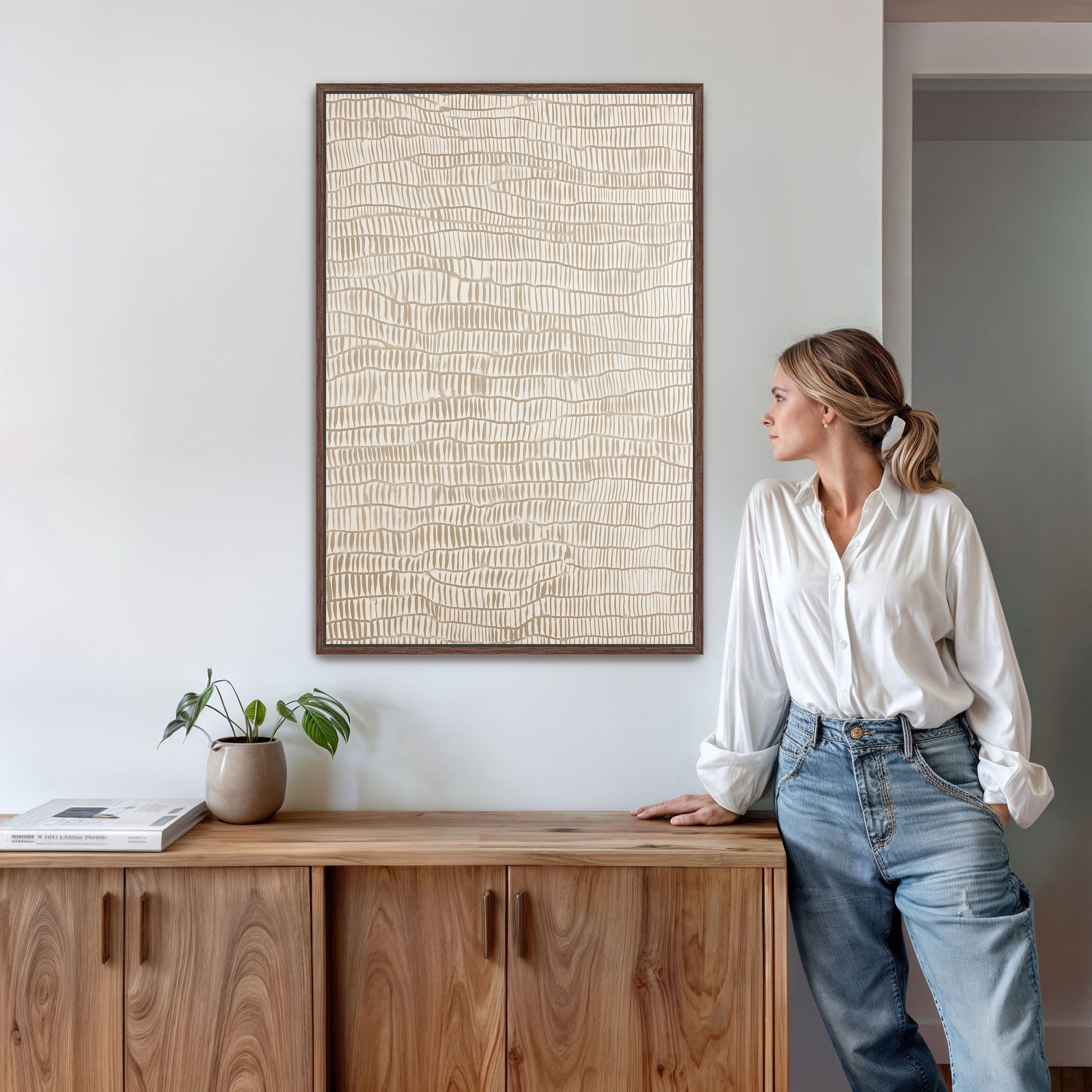 A woman in a white blouse and blue jeans stands next to a wooden cabinet, leaning against it with her hand on her hip. The cabinet has a plant on top of it, and there is a framed picture hanging on the wall above it.