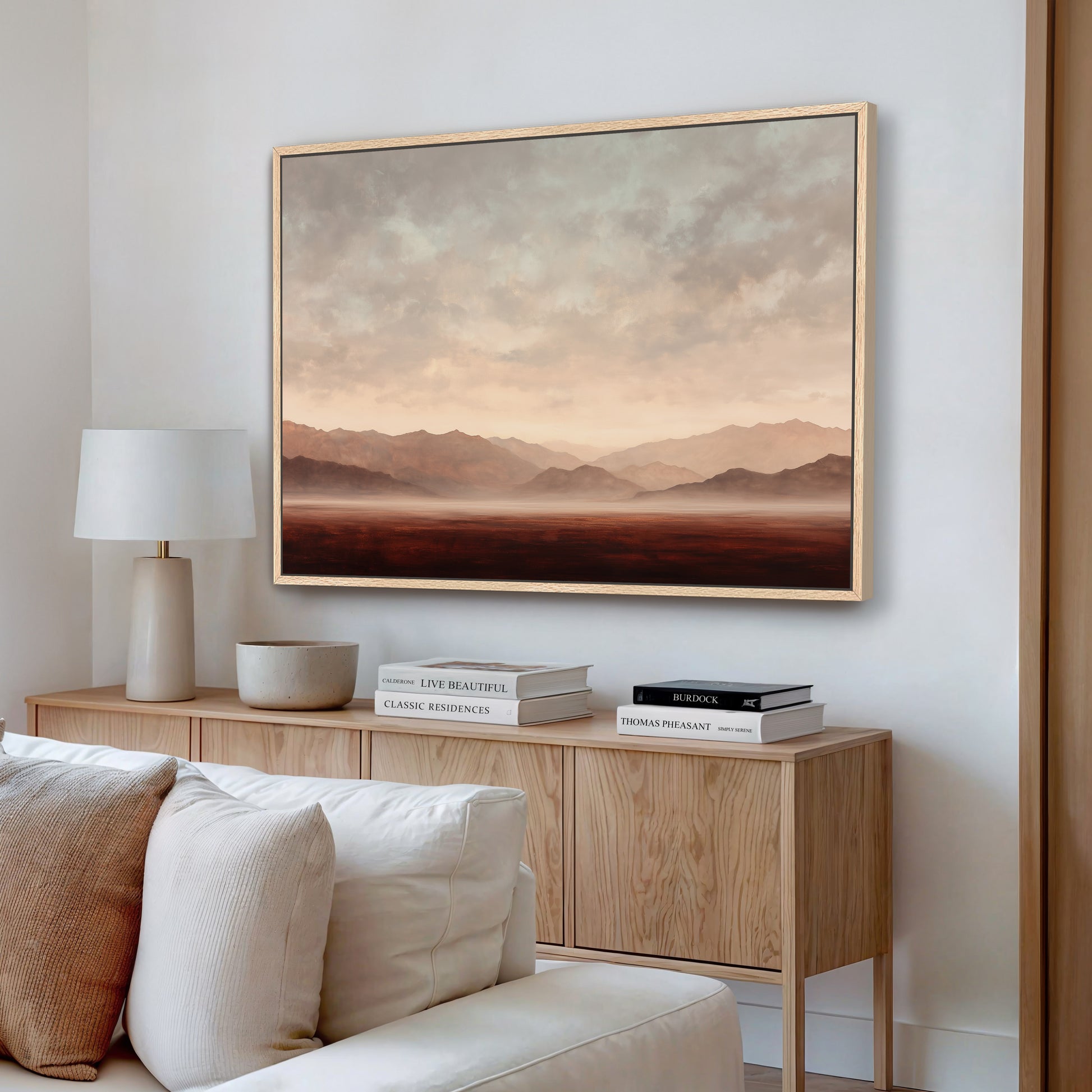 a living room with a large painting of a landscape hanging on the wall above a wooden cabinet. The cabinet has a bowl and a stack of books on it. The room also includes a white couch and a lamp.