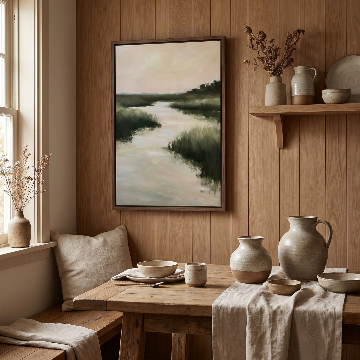 a cozy dining area with a wooden table set for two, featuring a vase, a bowl, and a pitcher. Above the table, there is a framed painting of a river and trees, adding a touch of nature to the space. The room has wooden paneling on the walls, and a window with a white curtain lets in natural light, creating a warm and inviting atmosphere.