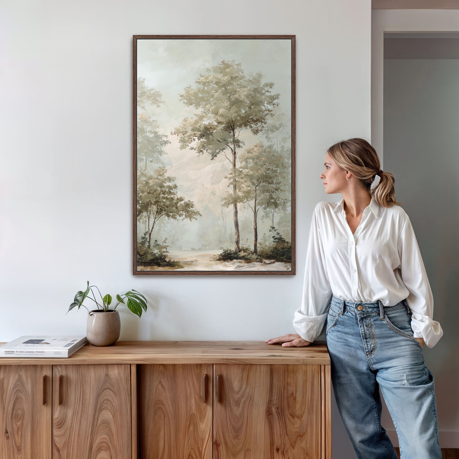 A woman in a white blouse and blue jeans stands in a room, leaning against a wooden cabinet, with a framed landscape painting hanging on the wall above her.