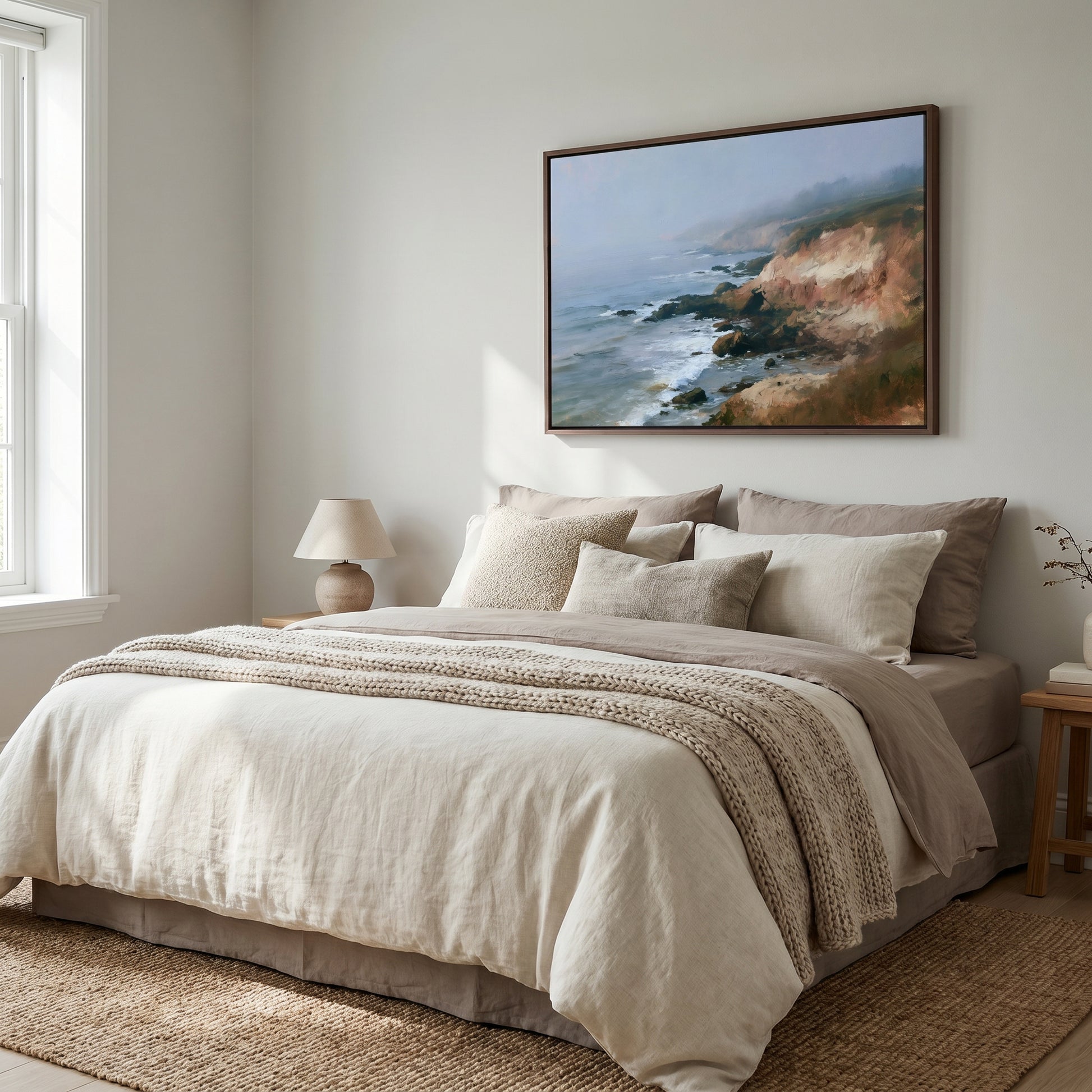 a serene bedroom scene with a large bed adorned with a white comforter and beige pillows, complemented by a wooden nightstand and a lamp. Above the bed, a framed painting of a coastal landscape adds a touch of nature and tranquility to the room.