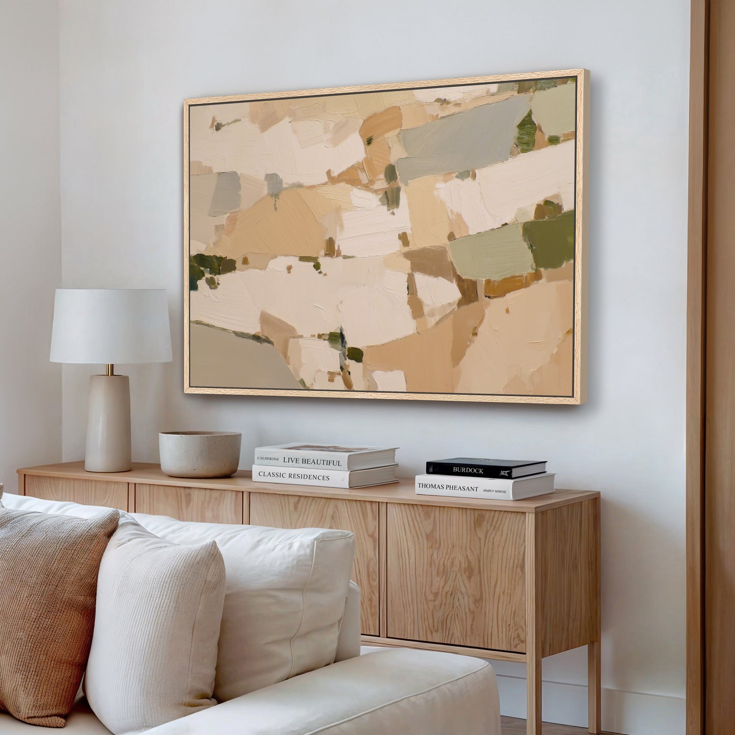 a living room with a large abstract painting hanging on the wall above a wooden console table. The console table has a lamp, a stack of books, and a bowl on it. The room has a white sofa and a wooden floor, creating a cozy and inviting atmosphere.