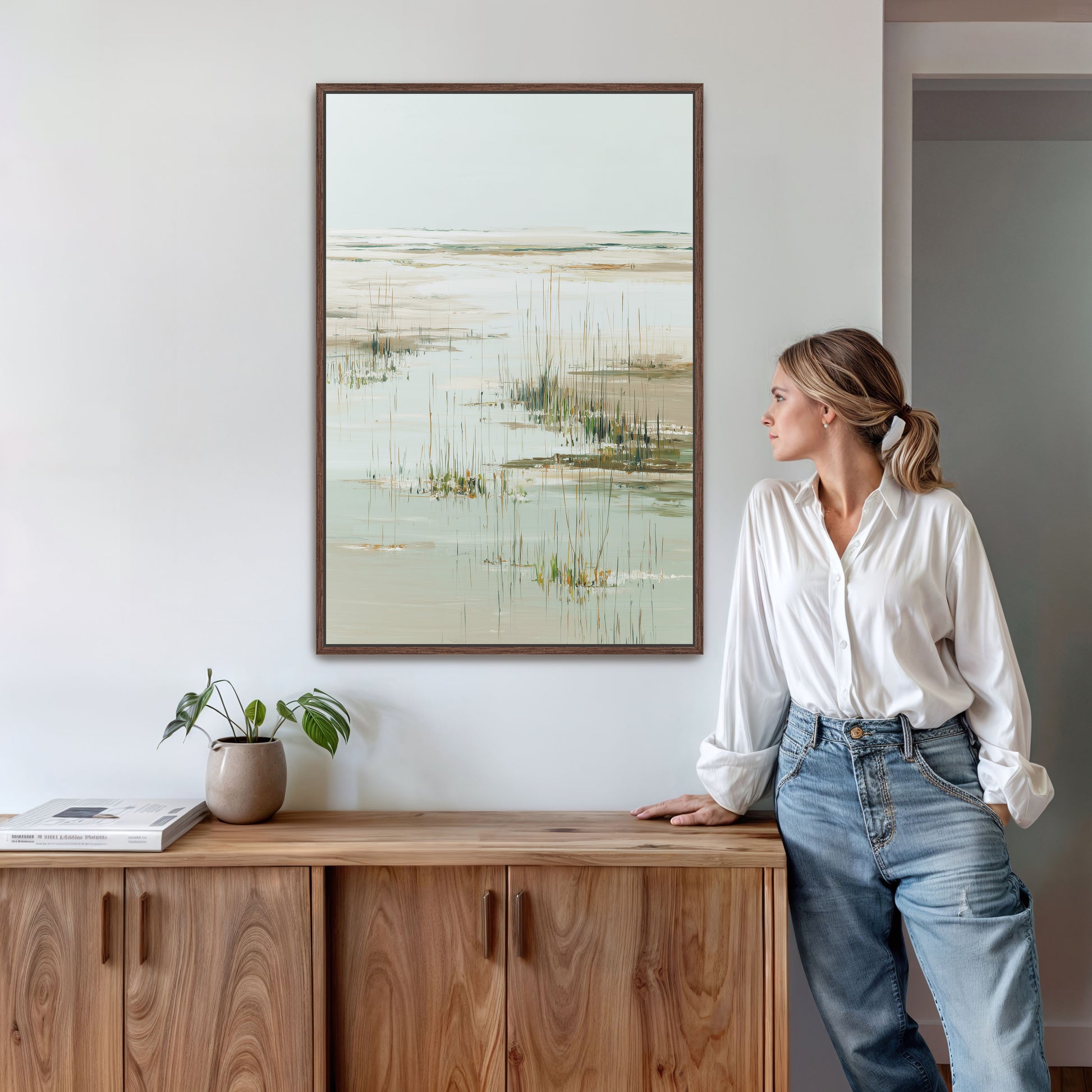 A woman in a white blouse and blue jeans stands in a room, leaning against a wooden cabinet, with a framed landscape painting hanging on the wall above her.