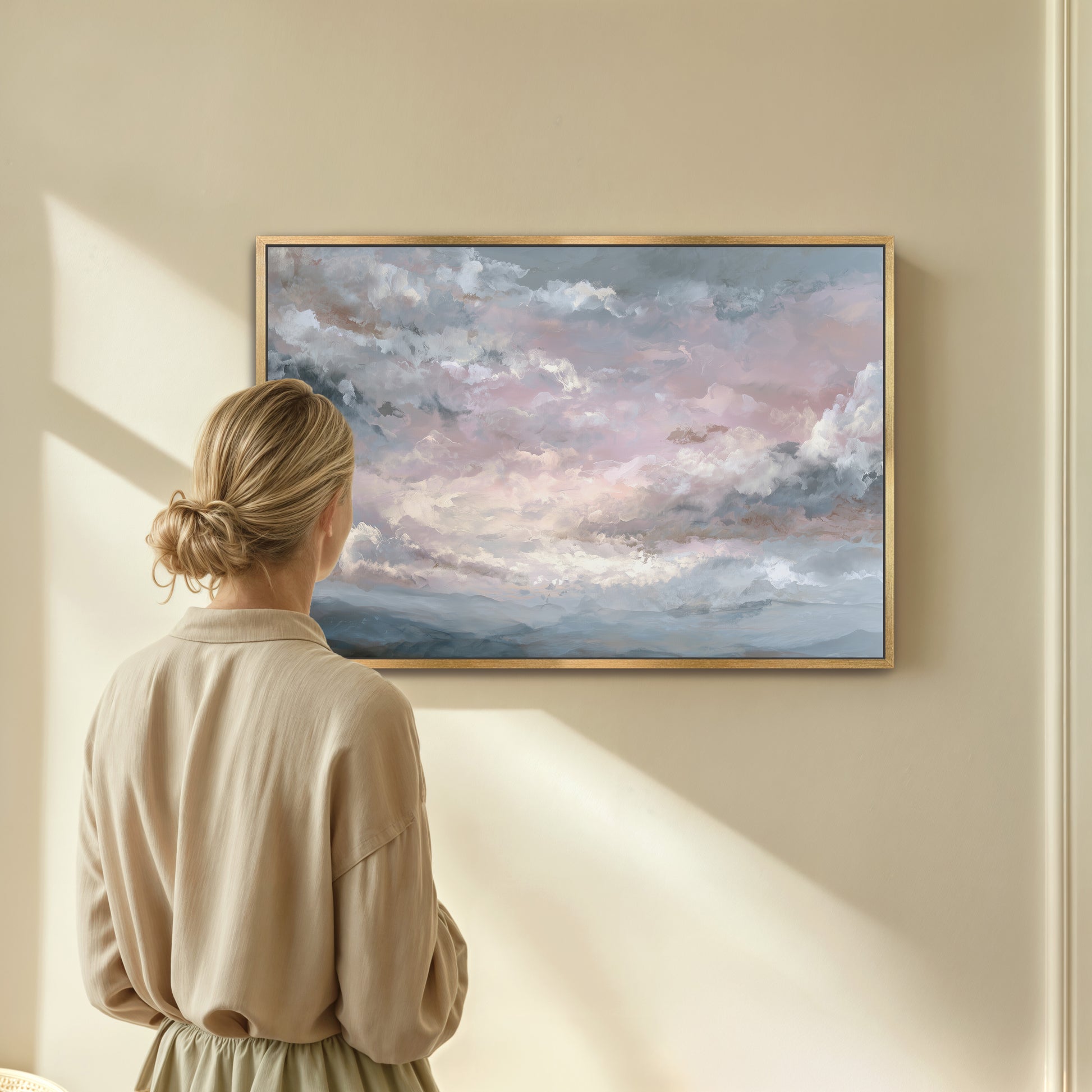 Woman admiring pastel sky artwork framed in light wood on beige wall.