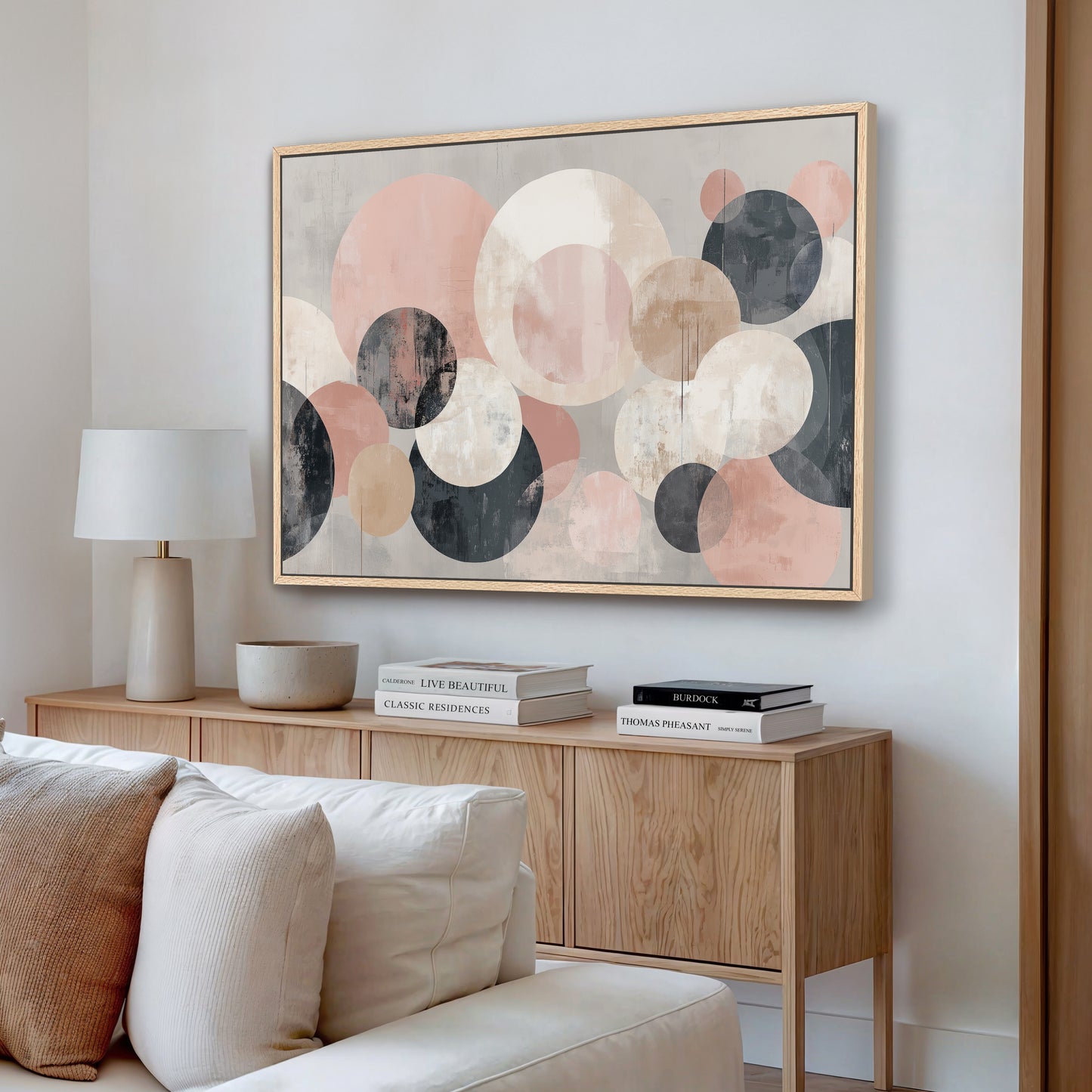 a living room with a large abstract painting hanging on the wall above a wooden cabinet. The cabinet has a bowl and a stack of books on it. The room also includes a white couch and a lamp.