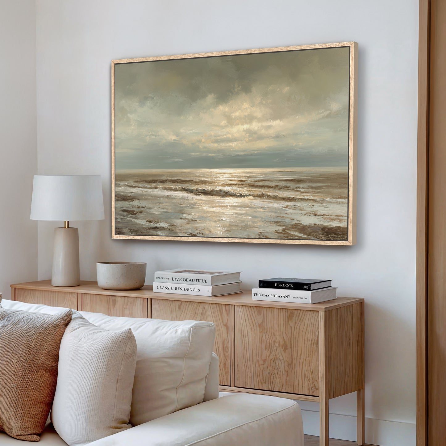 a living room with a large painting of a serene ocean landscape hanging on the wall above a wooden cabinet. The cabinet has a bowl and a stack of books on it. The room also includes a white sofa and a lamp, creating a cozy and inviting atmosphere.