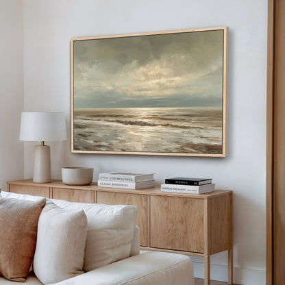 a living room with a large painting of a serene ocean landscape hanging on the wall above a wooden cabinet. The cabinet has a bowl and a stack of books on it. The room also includes a white sofa and a lamp, creating a cozy and inviting atmosphere.