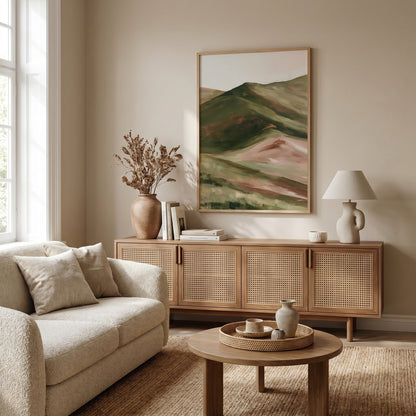 a cozy living room with a large painting of a mountain landscape hanging on the wall. The room is furnished with a beige sofa, a wooden sideboard, and a round coffee table. The room is illuminated by natural light coming through a window, creating a warm and inviting atmosphere.