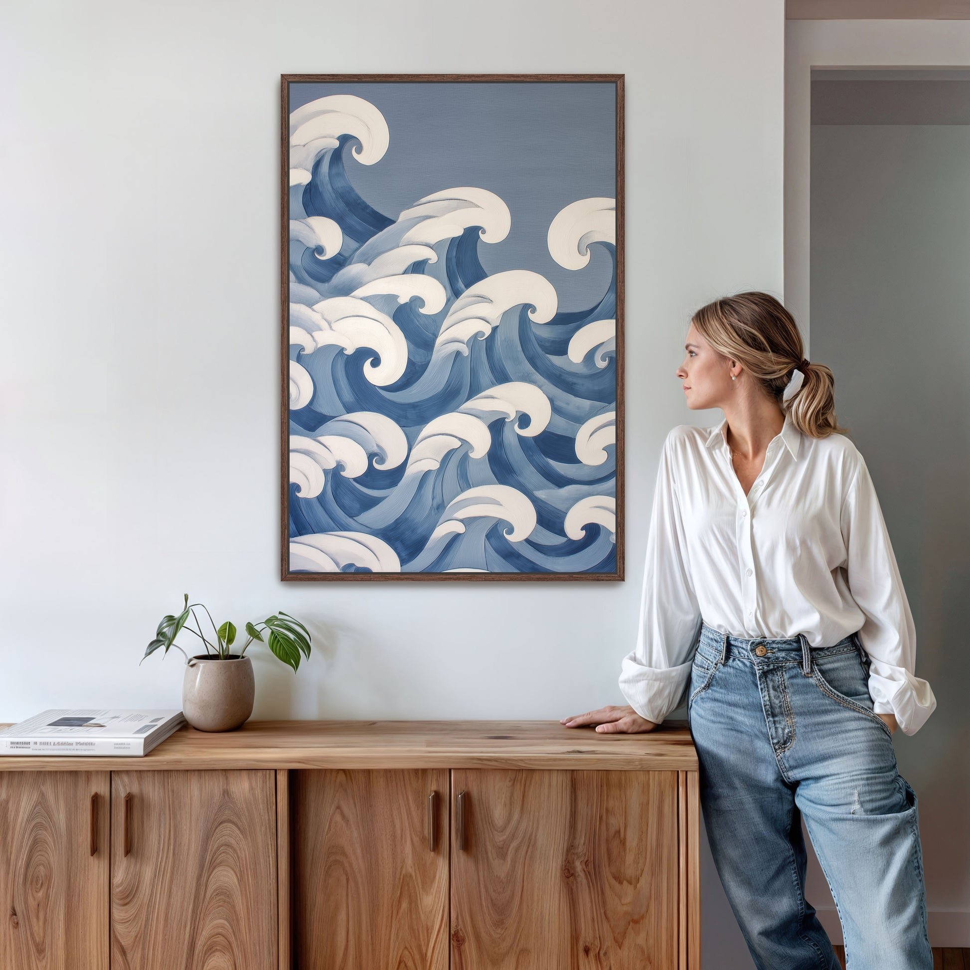 A woman stands in a room, leaning against a wooden cabinet, with a framed artwork of ocean waves hanging on the wall behind her.
